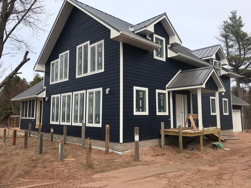 Blue-Build-White-Trim-1 A newly constructed two-story house with dark blue siding, a gray metal roof, and numerous white-framed windows, surrounded by red dirt and wooden posts.