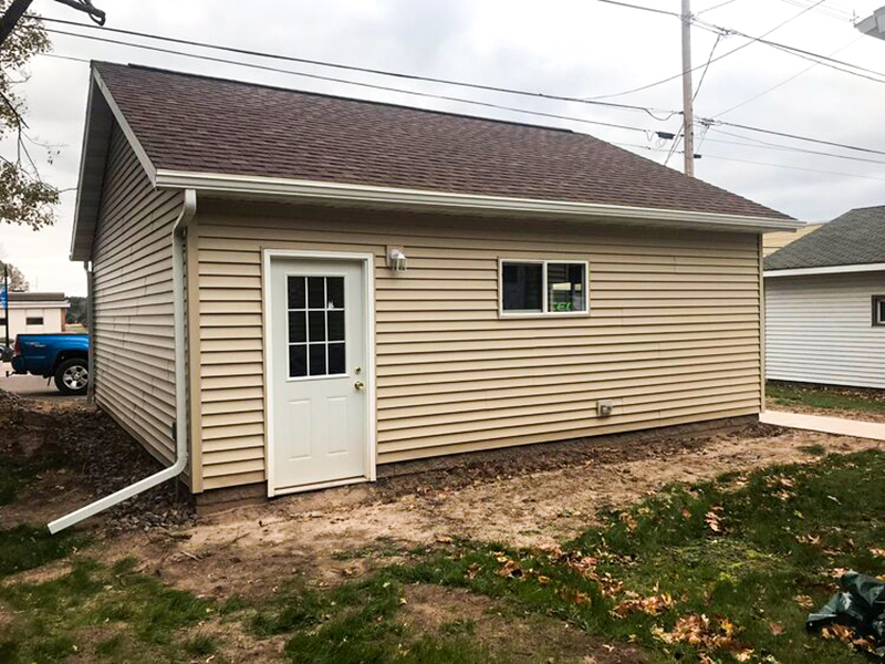 Detached-Garage-2 The side view of a newly built garage with beige siding, a brown roof, a white access door with a window, and a small white window.