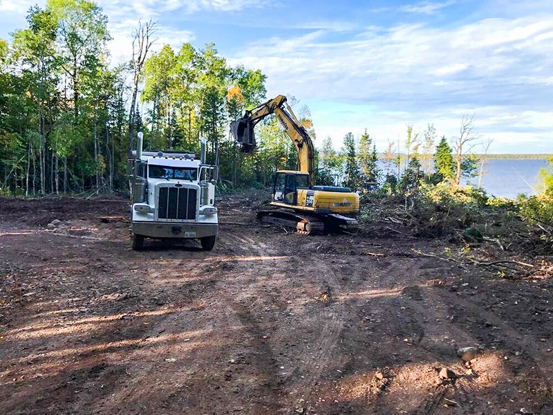 Excavator-Truck-2 A white dump truck and a yellow excavator are on a cleared dirt lot next to a forest and a body of water under a blue sky.