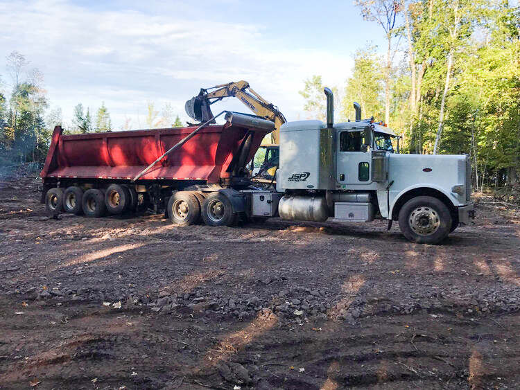 Excavator-Truck A white semi-truck is hitched to a large red dump trailer, and a yellow excavator is dumping material into it on a dirt lot.